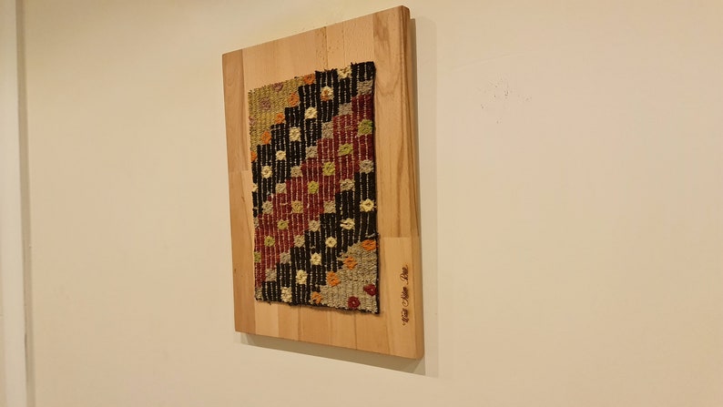 Puede incluir: Una alfombra tejida colorida con patrones geom&eacute;tricos en tonos rojos, negros, blancos y amarillos est&aacute; montada en una tabla de madera. La tabla tiene el texto "Hand Woven Rug" inscrito en ella.