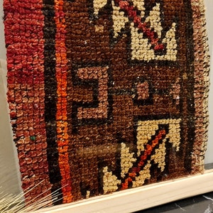 May include: A framed piece of vintage fabric with a colorful geometric pattern in shades of red, orange, brown, and white. The fabric is framed in a natural wood frame.