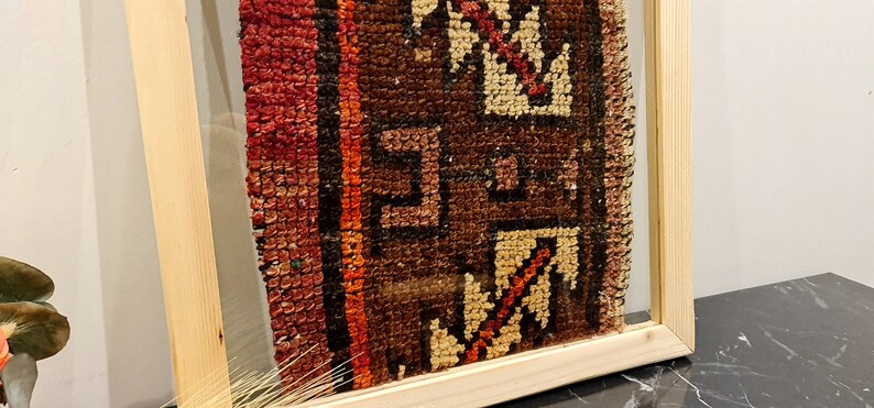 May include: A framed piece of vintage fabric with a colorful geometric pattern in shades of red, orange, brown, and white. The fabric is framed in a natural wood frame.