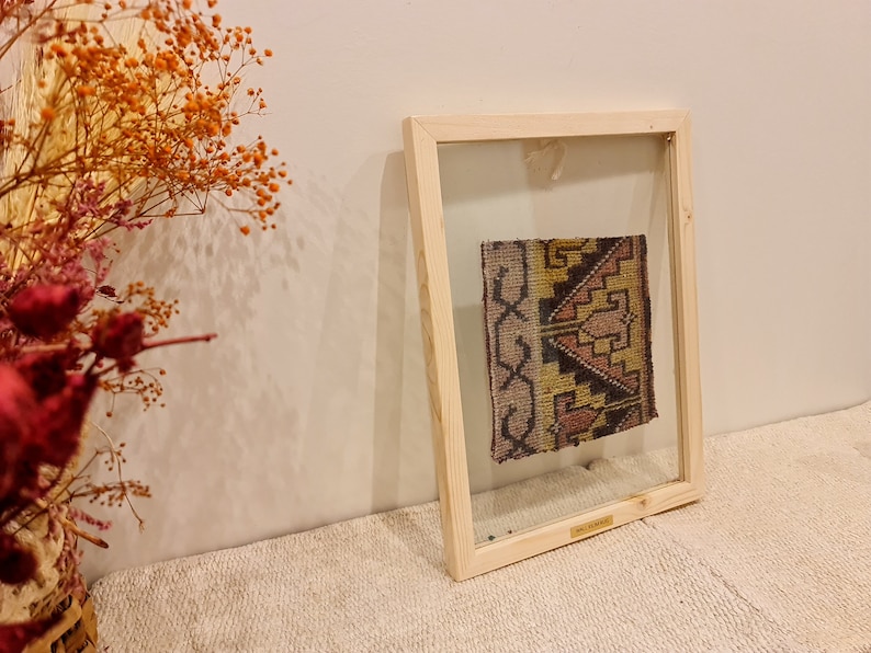 May include: A framed piece of colorful woven fabric with geometric patterns in shades of pink, yellow, and brown. The fabric is displayed in a clear glass frame with a wooden border.