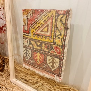 May include: A framed vintage rug with a colourful geometric pattern in shades of red, yellow, brown, and white. The rug has a fringed edge and is displayed in a simple wooden frame.
