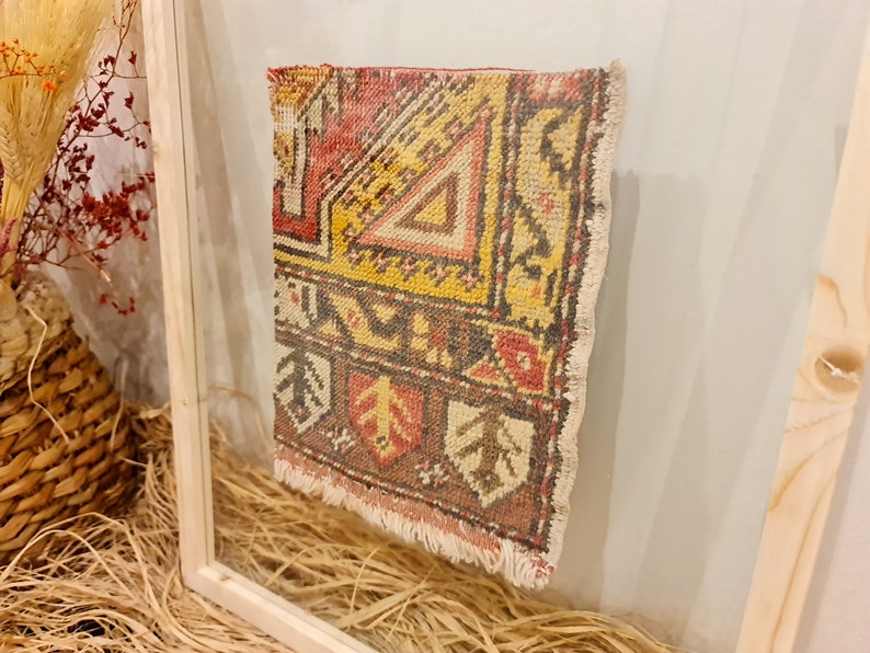 May include: A framed vintage rug with a colourful geometric pattern in shades of red, yellow, brown, and white. The rug has a fringed edge and is displayed in a simple wooden frame.