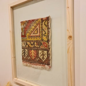 May include: A framed piece of colourful, patterned fabric with a fringe edge. The fabric is a traditional rug design with geometric shapes in shades of red, yellow, brown, and white.