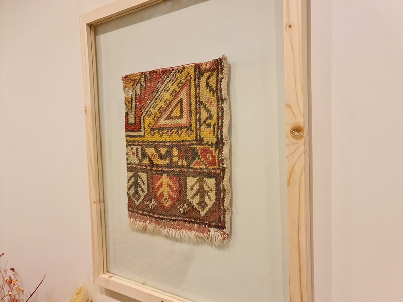 May include: A framed piece of colourful, patterned fabric with a fringe edge. The fabric is a traditional rug design with geometric shapes in shades of red, yellow, brown, and white.