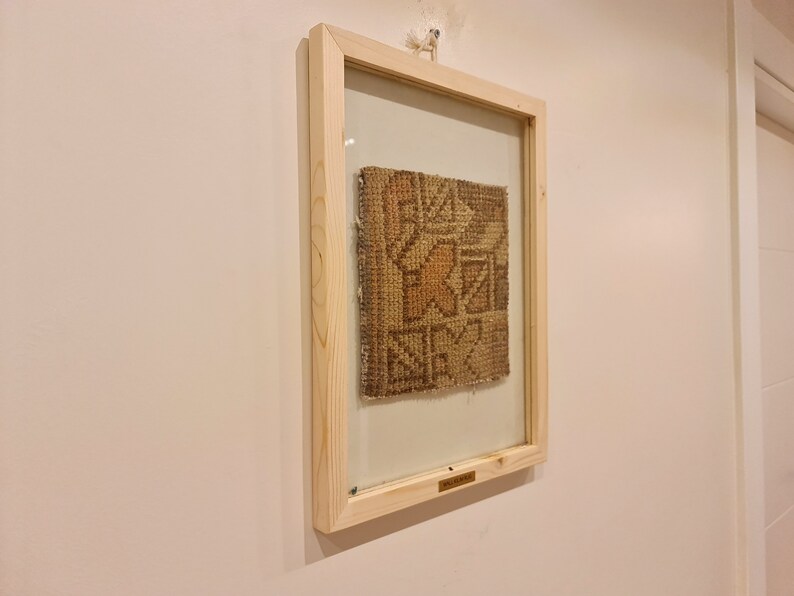 May include: A framed piece of woven fabric with a geometric pattern in shades of brown and beige. The frame is made of natural wood and has a glass front.