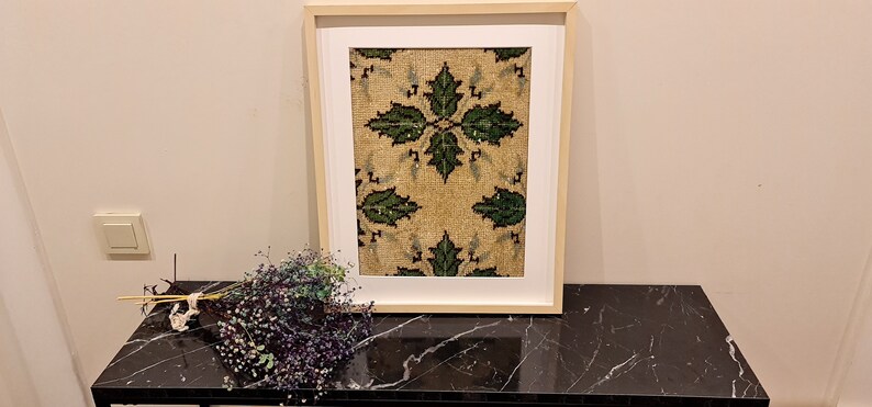May include: A framed artwork with a beige background and a repeating pattern of green leaves. The frame is made of wood and has a white mount.