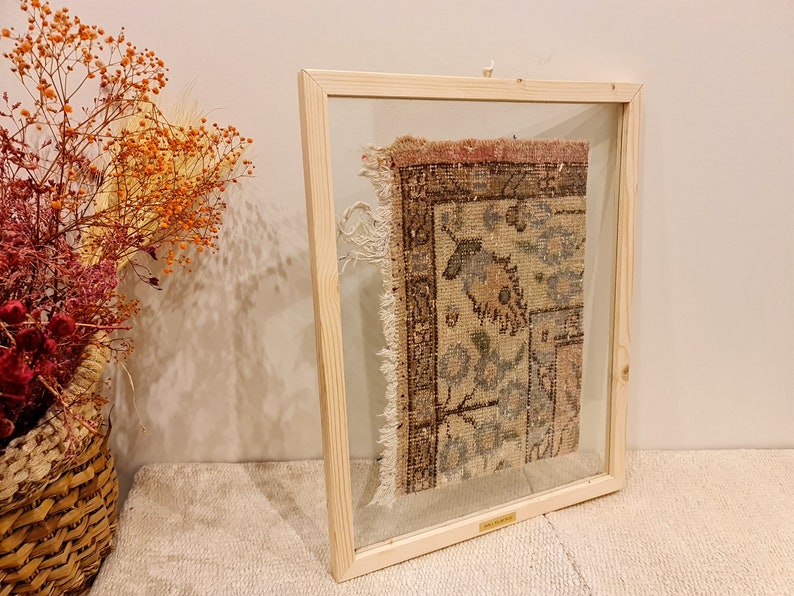May include: A framed vintage rug with a faded floral pattern in shades of pink, blue, and brown. The rug is displayed in a simple wooden frame with a glass front.