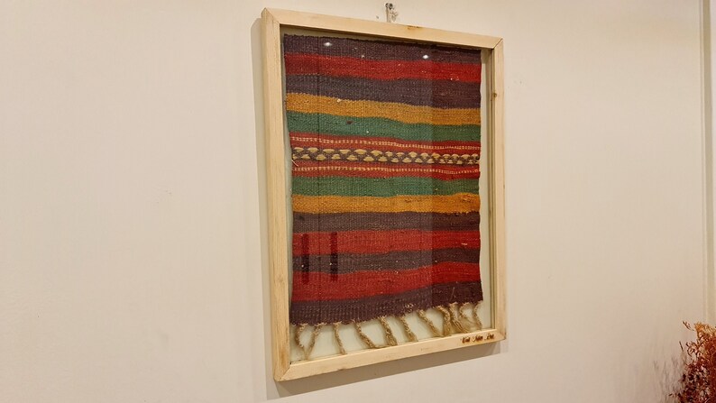 May include: A framed tapestry with a colorful striped pattern in shades of red, brown, green, and yellow. The tapestry has a fringe at the bottom.