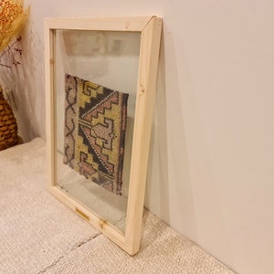 May include: A framed piece of colorful fabric with geometric patterns in shades of pink, yellow, and black. The fabric is displayed in a simple wooden frame with a clear glass front.