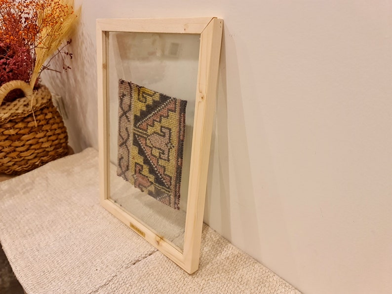 May include: A framed piece of colorful fabric with geometric patterns in shades of pink, yellow, and black. The fabric is displayed in a simple wooden frame with a clear glass front.