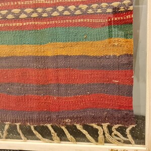May include: A framed woven tapestry with a colorful striped pattern in shades of red, green, yellow, and purple. The tapestry has a fringe at the bottom.