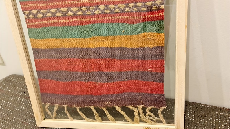May include: A framed woven tapestry with a colorful striped pattern in shades of red, green, yellow, and purple. The tapestry has a fringe at the bottom.