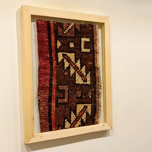 May include: A framed piece of fabric with a geometric pattern in shades of brown, red, and cream. The fabric is framed in a natural wood frame.