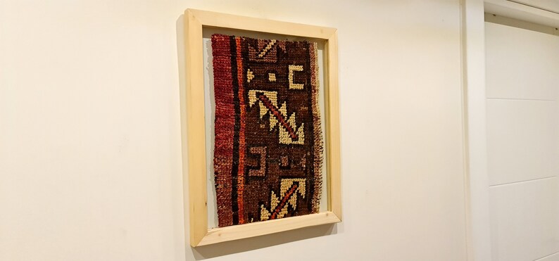 May include: A framed piece of fabric with a geometric pattern in shades of brown, red, and cream. The fabric is framed in a natural wood frame.