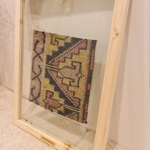 May include: A framed small kilim rug with a geometric pattern in shades of pink, yellow, black, and gray. The frame is made of natural wood and has a glass front. The label on the bottom of the frame reads "Small Kilim Rug".
