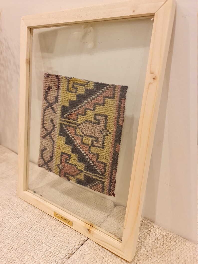 May include: A framed small kilim rug with a geometric pattern in shades of pink, yellow, black, and gray. The frame is made of natural wood and has a glass front. The label on the bottom of the frame reads "Small Kilim Rug".
