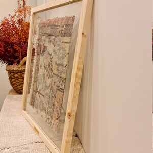 May include: A framed piece of fabric with a colorful pattern, displayed on a white wall. The frame is made of natural wood and has a glass front.