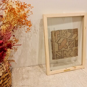 May include: A framed piece of woven fabric with a geometric pattern in shades of brown, beige, and pink. The frame is made of light wood and has a clear glass front.