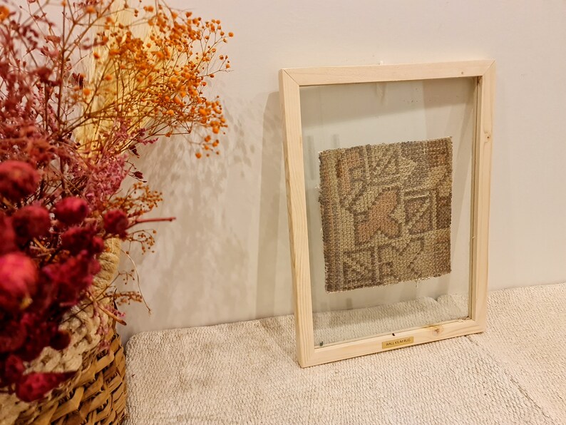 May include: A framed piece of woven fabric with a geometric pattern in shades of brown, beige, and pink. The frame is made of light wood and has a clear glass front.