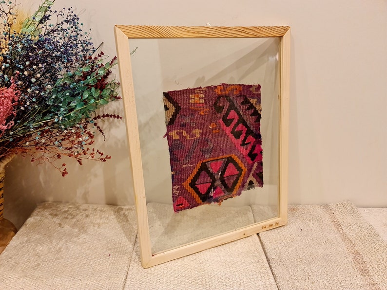 May include: A framed piece of colorful fabric with a geometric pattern. The fabric is a mix of pink, purple, orange, and black. The frame is made of light wood and has a clear glass front.
