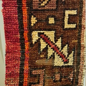 May include: A close-up of a framed rug with a geometric pattern in shades of red, brown, and beige. The rug is woven with intricate details and has a vintage look.