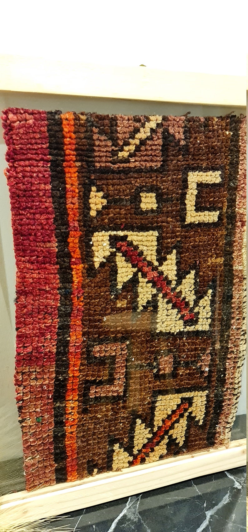 May include: A close-up of a framed rug with a geometric pattern in shades of red, brown, and beige. The rug is woven with intricate details and has a vintage look.