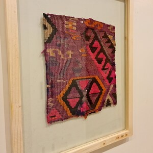 May include: A framed piece of colorful woven fabric with a geometric pattern in shades of pink, orange, black, and purple.
