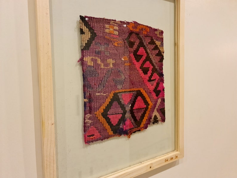 May include: A framed piece of colorful woven fabric with a geometric pattern in shades of pink, orange, black, and purple.