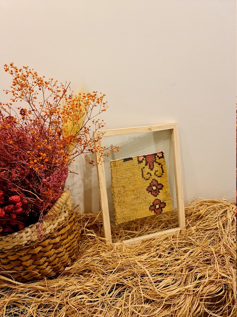 Puede incluir: Un trozo de tela amarilla enmarcado con un patr&oacute;n floral rojo y negro, exhibido sobre un lecho de material similar a la paja. Un ramo de flores secas en una cesta de mimbre est&aacute; en primer plano.