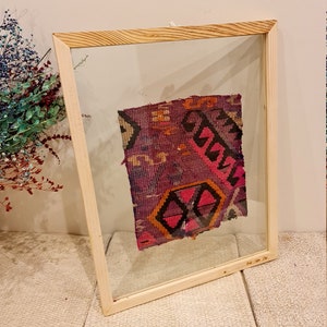 May include: A framed piece of colorful fabric with a geometric pattern in shades of pink, purple, orange, and black. The fabric is displayed in a wooden frame with a clear glass front.