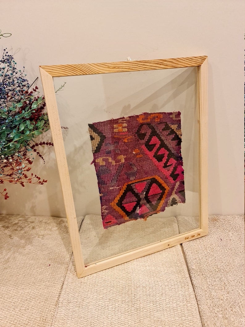 May include: A framed piece of colorful fabric with a geometric pattern in shades of pink, purple, orange, and black. The fabric is displayed in a wooden frame with a clear glass front.