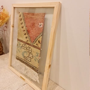 May include: A framed vintage rug with a pink, green and yellow geometric pattern. The rug is framed in a natural wood frame with a clear glass front.