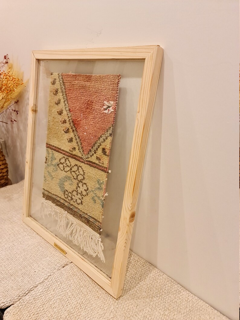 May include: A framed vintage rug with a pink, green and yellow geometric pattern. The rug is framed in a natural wood frame with a clear glass front.