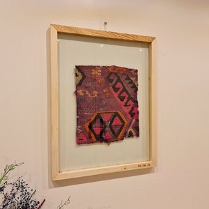 May include: A framed piece of fabric with a colorful geometric pattern in shades of pink, purple, orange, and black. The fabric is framed in a natural wood frame with a glass front.