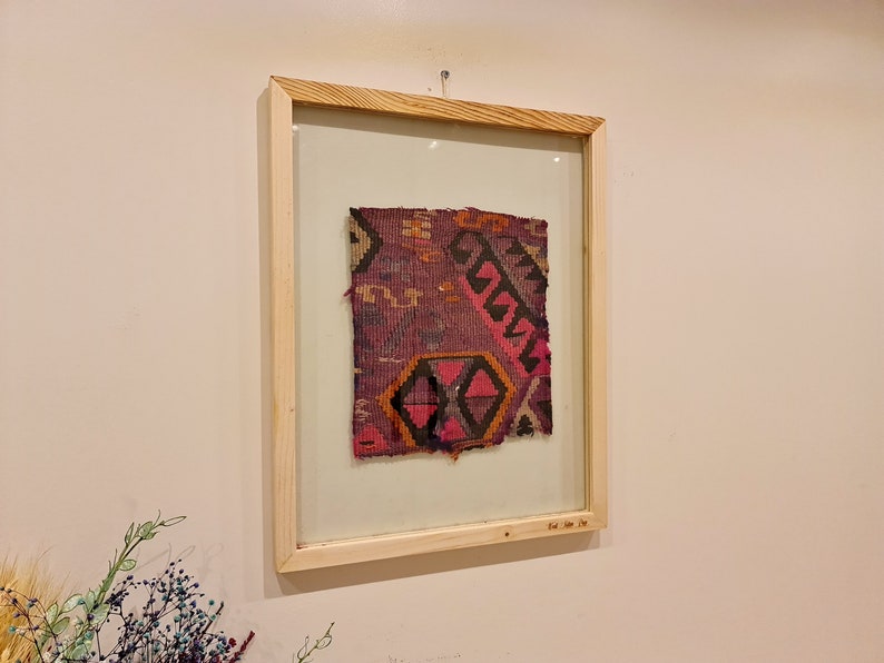 May include: A framed piece of fabric with a colorful geometric pattern in shades of pink, purple, orange, and black. The fabric is framed in a natural wood frame with a glass front.