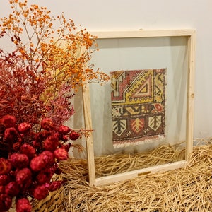 May include: A framed piece of colourful patterned fabric with a wooden frame and a clear glass front. The frame is sitting on a bed of straw.