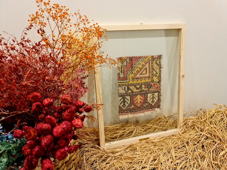 May include: A framed piece of colourful patterned fabric with a wooden frame and a clear glass front. The frame is sitting on a bed of straw.