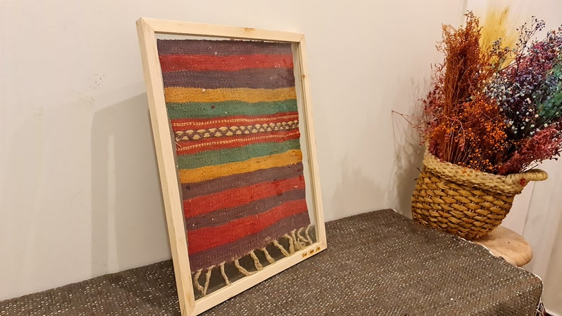 May include: A framed piece of colorful woven fabric with a wood frame. The fabric has stripes of red, purple, yellow, green, and brown.
