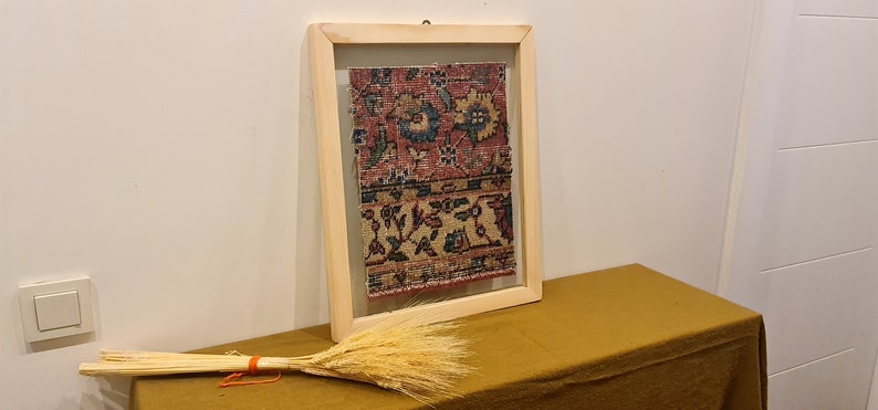 Pu&ograve; includere: Un pezzo di tessuto antico incorniciato con un motivo floreale colorato in tonalit&agrave; di rosso, blu e giallo. Il tessuto &egrave; esposto su una cornice in legno con una finitura marrone chiaro. La cornice &egrave; appoggiata su un tavolo marrone.