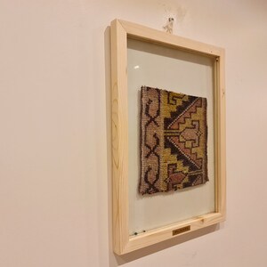 May include: A framed piece of fabric with a geometric pattern in shades of brown, yellow, pink, and white. The fabric is displayed in a wooden frame with a glass front.