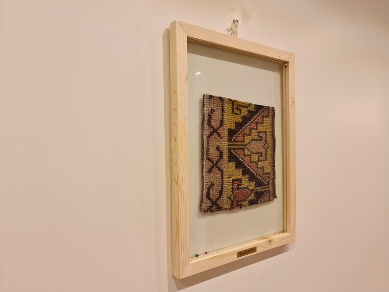 May include: A framed piece of fabric with a geometric pattern in shades of brown, yellow, pink, and white. The fabric is displayed in a wooden frame with a glass front.