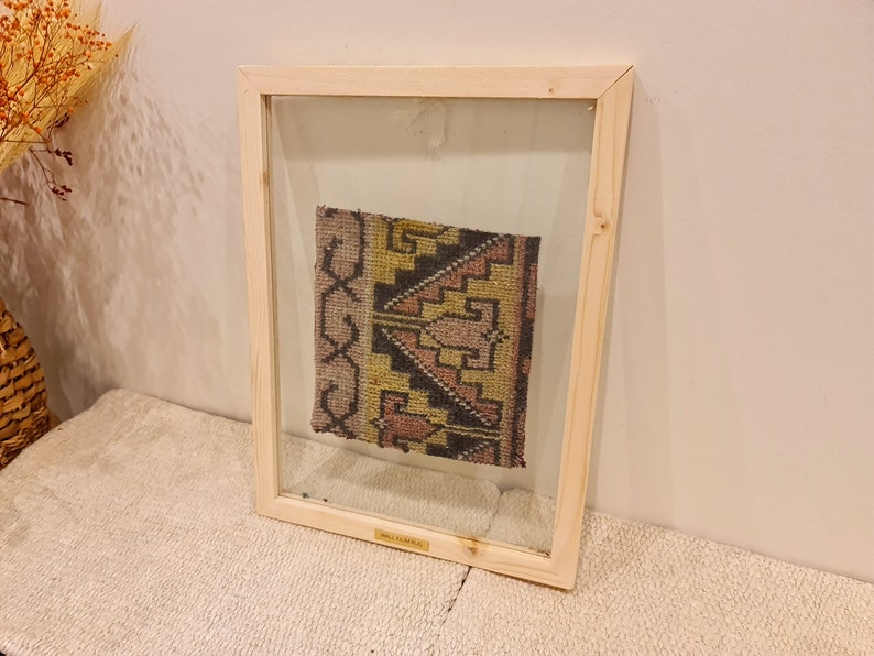May include: A framed piece of fabric with a geometric pattern in shades of pink, yellow, and black. The fabric is displayed in a wooden frame with a clear glass front.