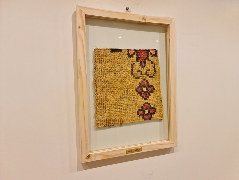 Puede incluir: Una alfombra de pared enmarcada con fondo amarillo y motivos florales rojos y negros. El marco es de madera clara y tiene una peque&ntilde;a etiqueta que dice "Alfombra de pared".