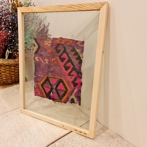 May include: A framed piece of colorful woven fabric with a geometric pattern in shades of pink, purple, black, and orange. The frame is made of natural wood and has a glass front. The text "East Anatolia 19th" is inscribed on the bottom of the frame.