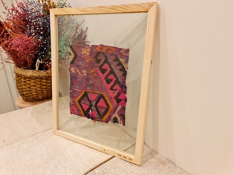 May include: A framed piece of colorful woven fabric with a geometric pattern in shades of pink, purple, black, and orange. The frame is made of natural wood and has a glass front. The text "East Anatolia 19th" is inscribed on the bottom of the frame.