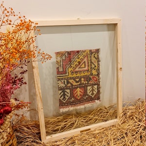 May include: A framed vintage rug with a colourful geometric pattern in shades of red, yellow, and brown. The rug is displayed in a wooden frame with a clear glass front.