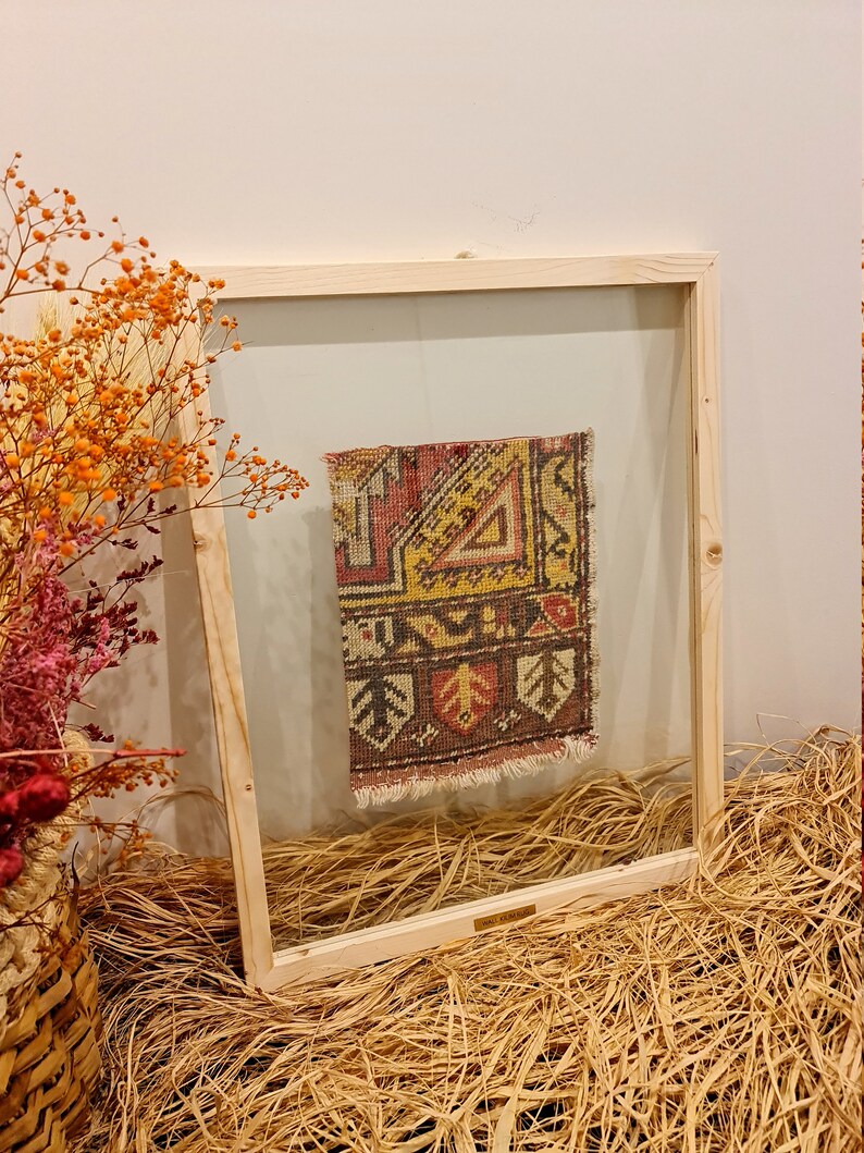 May include: A framed vintage rug with a colourful geometric pattern in shades of red, yellow, and brown. The rug is displayed in a wooden frame with a clear glass front.
