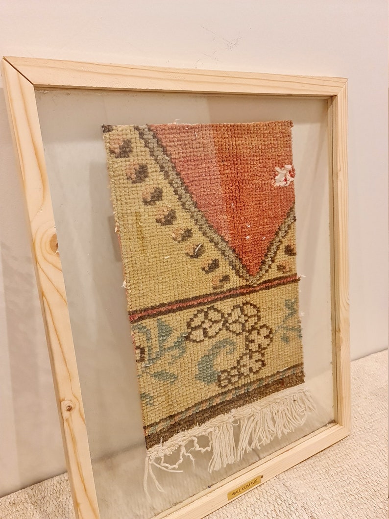 May include: A framed vintage rug with a cream, pink and brown geometric pattern. The rug has fringe on the bottom and is labelled "Wall Rug".