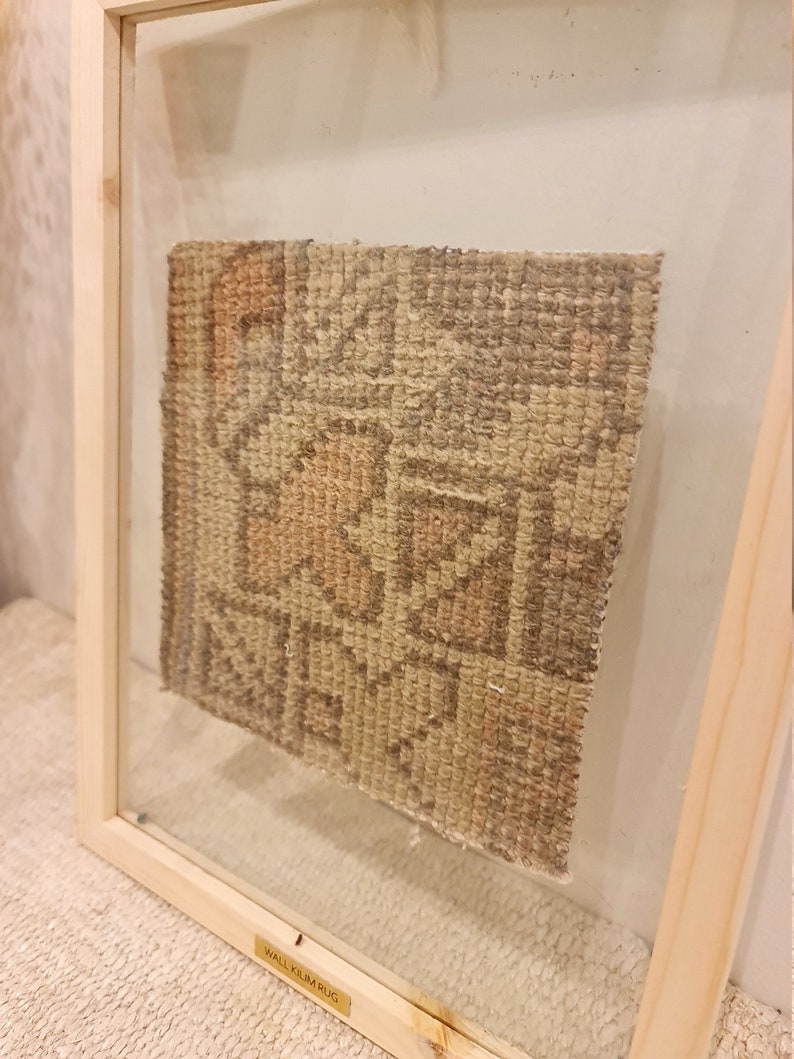 May include: A framed beige and brown woven wall kilim rug with a geometric pattern. The frame is made of natural wood and has a glass front. The label on the frame reads "Wall Kilim Rug".