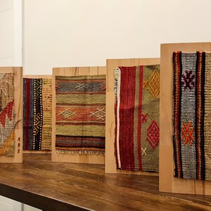 May include: Four framed samples of colorful woven fabric with intricate patterns. The fabric is mounted on wooden boards and displayed on a wooden table.